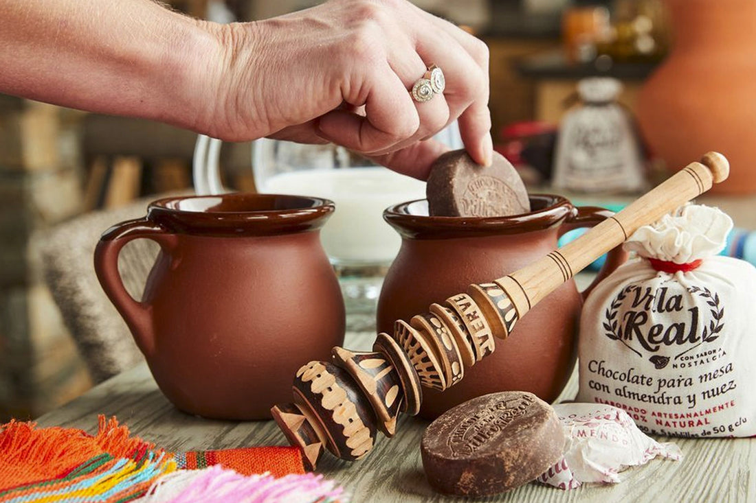 Hand placing a disc of Mexican drinking chocolate into a traditional clay mug, with a wooden molinillo, Villa Real chocolate packaging, and colorful textiles on a rustic table.