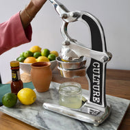 Hand-operated metal citrus juicer with black grip handle on marble base, collecting fresh lemon juice in glass jar.