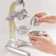 Silver metal citrus juicer with lever handle, cream-colored insert panel, and two metal cups being held by hands.