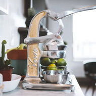 Manual citrus juicer with silver metal frame, wooden handle, and stainless steel collection bowls.