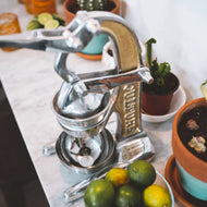 Manual citrus juicer with silver metal lever and brass-colored band, positioned on white surface with fresh citrus fruits nea