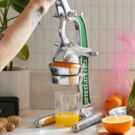 Hand-operated citrus juicer with metal lever and green handle positioned over glass of fresh juice, with orange half and ging
