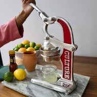 Hand-operated citrus juicer with red handle and metal frame on marble base, with lemons and limes nearby.