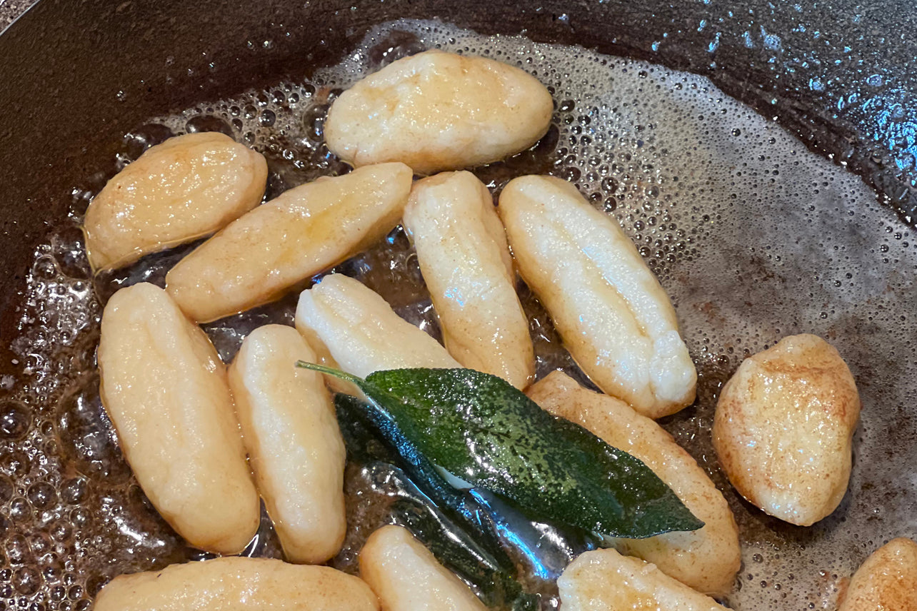 Gnocchi and garganelli pasta cooking in boiling water with sage leaf and butter in dark pan.