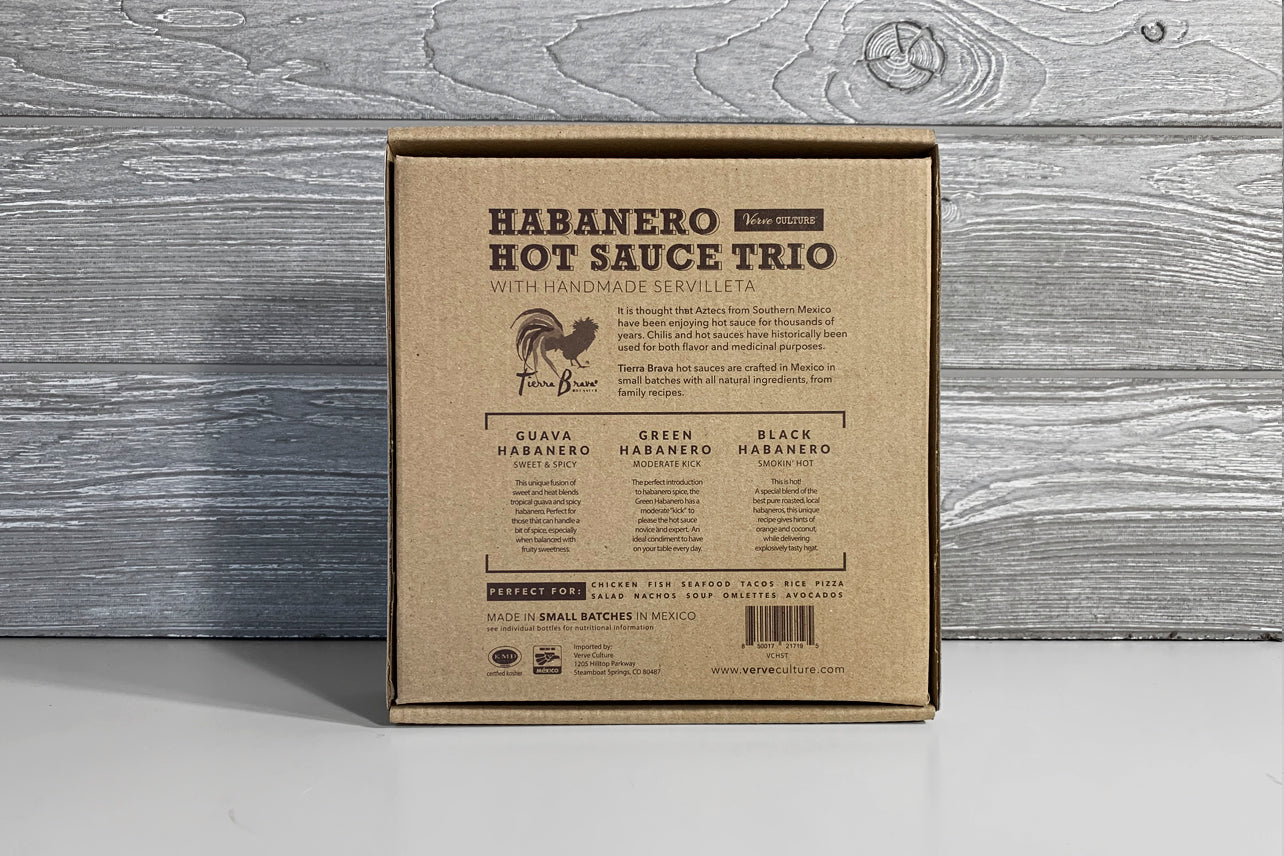 Cardboard box containing habanero hot sauce trio and handmade servilleta on wooden surface.