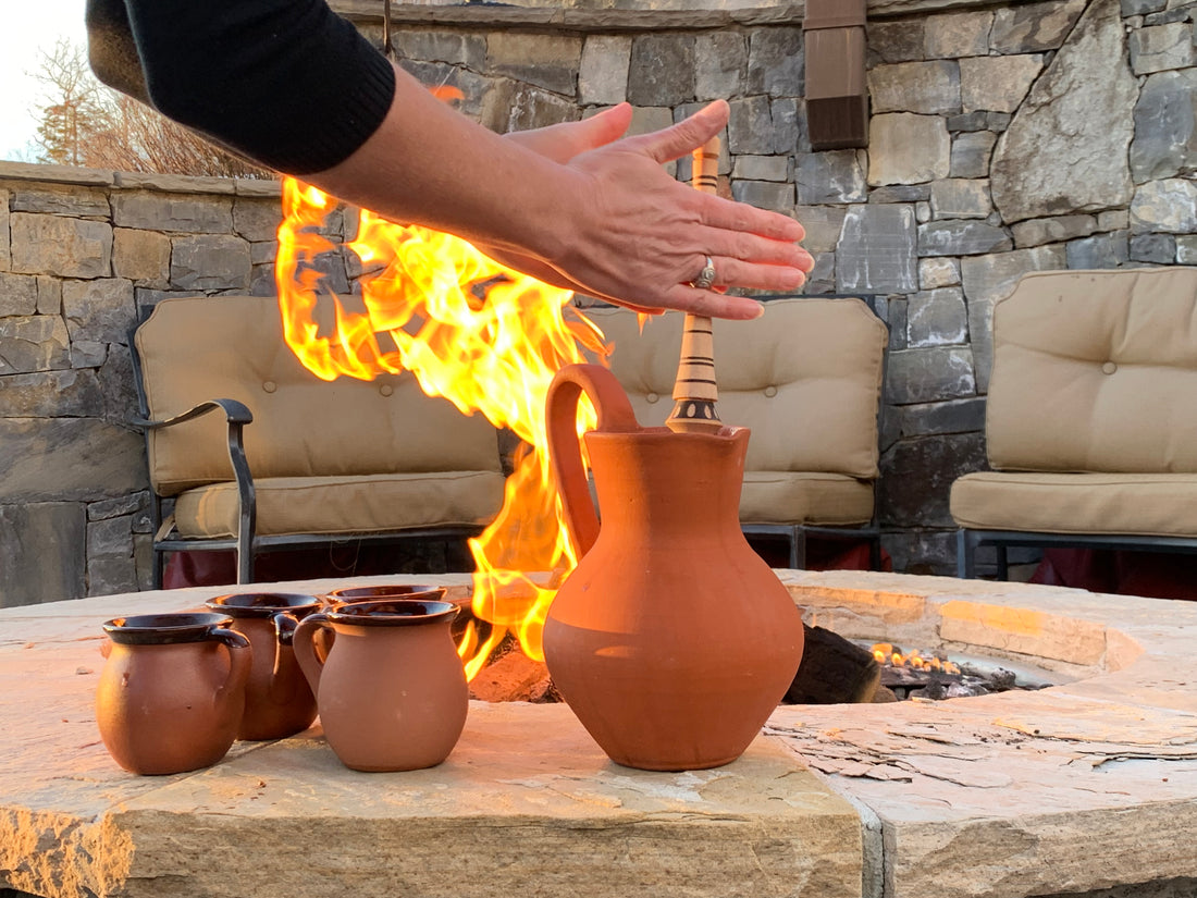 Hand warming over a fire pit beside traditional clay pitchers and mugs outdoors, with a stone wall and patio seating in the background