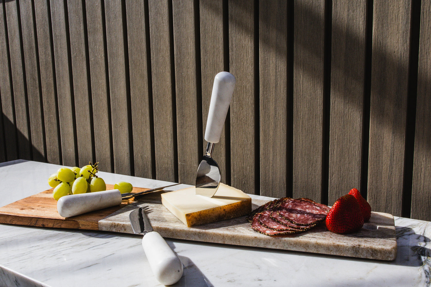 Three cheese knives with white marble handles displayed on marble boards with cheese, cured meat, grapes, and strawberries.