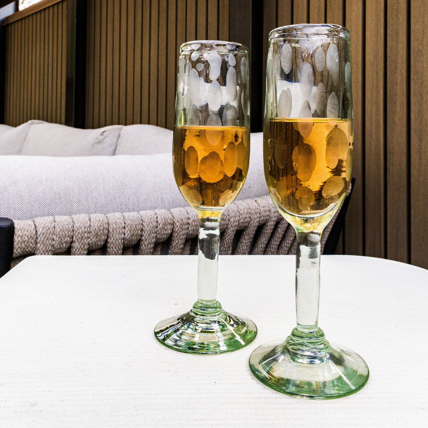 Two clear glass champagne flutes with etched circular patterns, filled with golden liquid, featuring green-tinted bases.