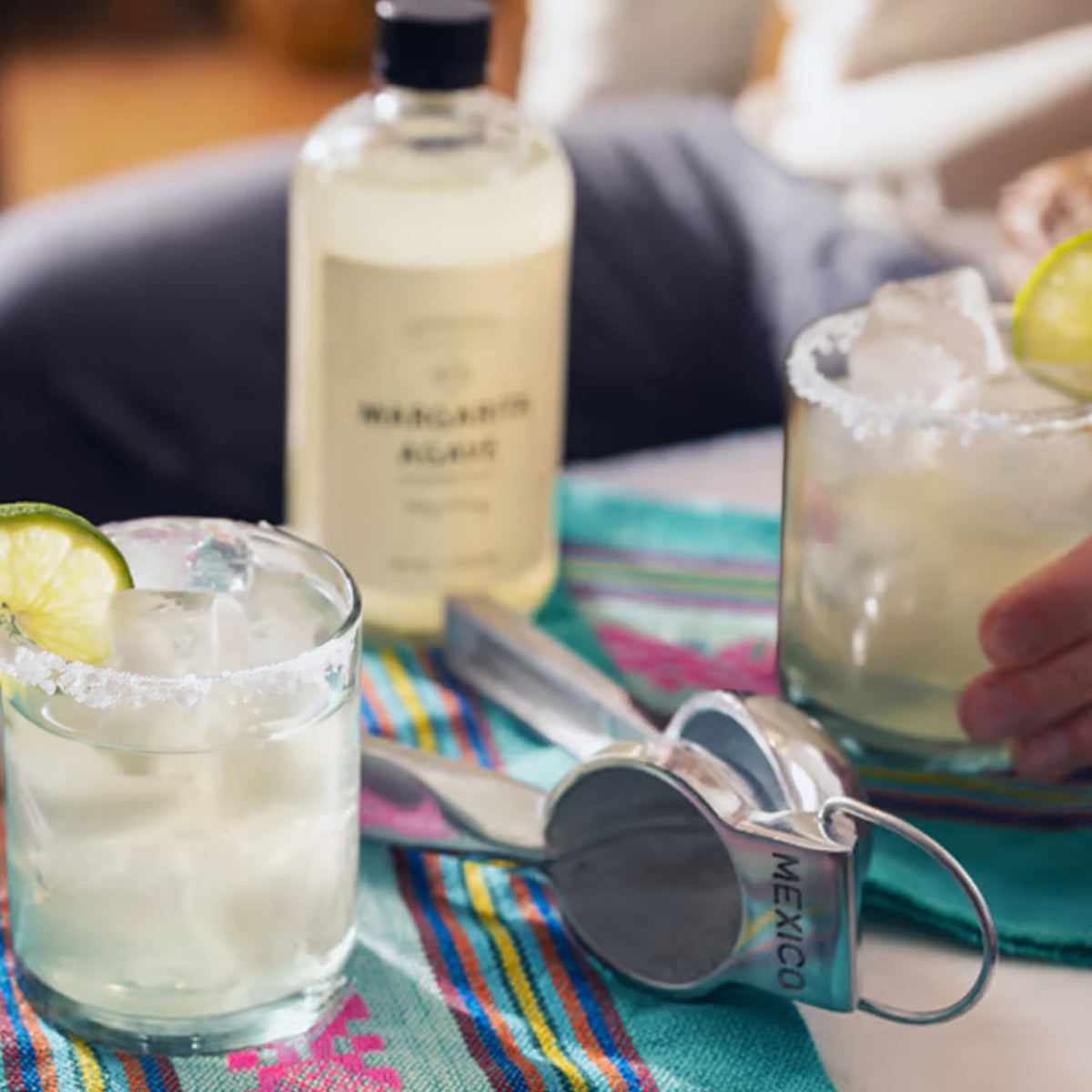Clear cocktails with lime slices and salt rims, a bottle of margarita mix, measuring jigger, and colorful striped napkins arr