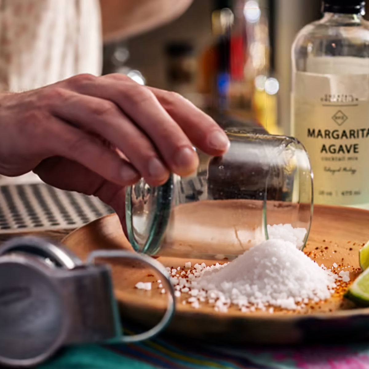 Hand pouring salt from glass onto wooden surface with margarita agave bottle and cocktail ingredients in background.