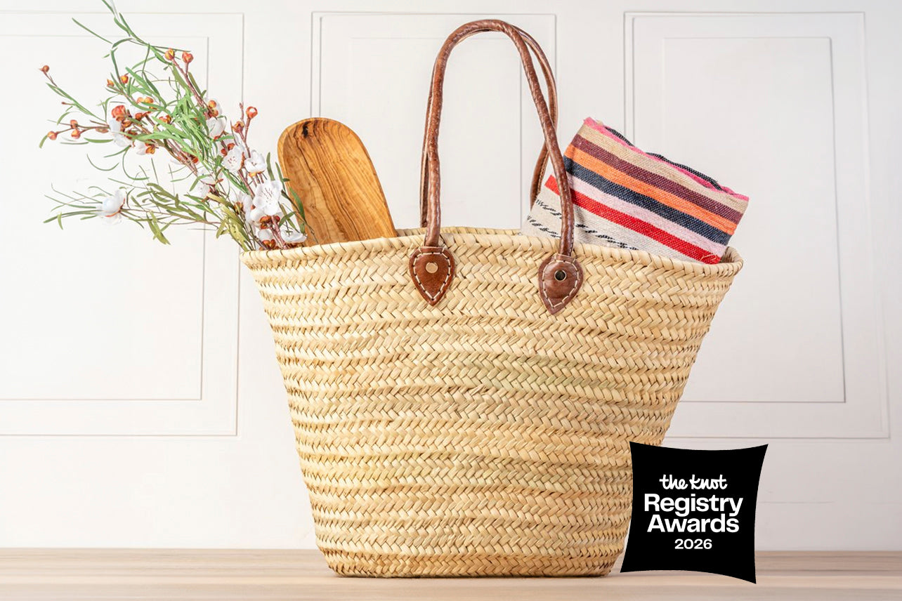 Woven straw tote basket with brown leather handles containing dried flowers, wooden spoon, and striped fabric.